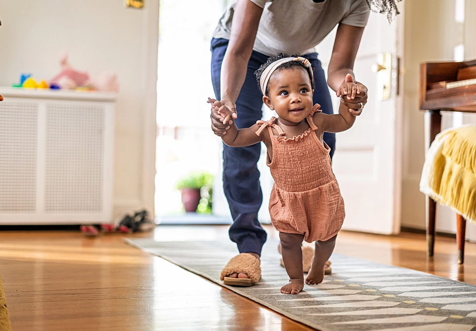 A baby taking their first steps, supported by a parent's hands.